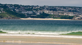 Carbis Bay frrom Hayle This is a landscape photograph showing Carbis Bay from Hayle in Cornwall, captured in the late morning during early summer. The image features the sea in the foreground, with gentle waves rolling onto the sandy beach. In the distance, the town of St Ives is visible, with its distinctive array of houses and buildings spread across the hillside. The scene captures the coastal beauty of Cornwall, highlighting the relationship between the sea, the expansive beach, and the urban area of St Ives.